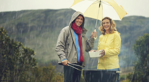 man and woman stood underneath and umbrella cooking on a BBQ