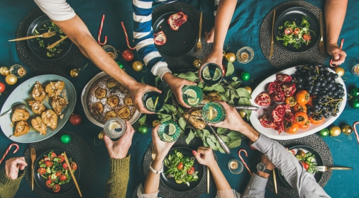 Company of friends of different ages or family gathering for Christmas or New Year party dinner at festive table. Flat-lay of human hands holding glasses with drinks, feasting and celebrating holiday together, top view.